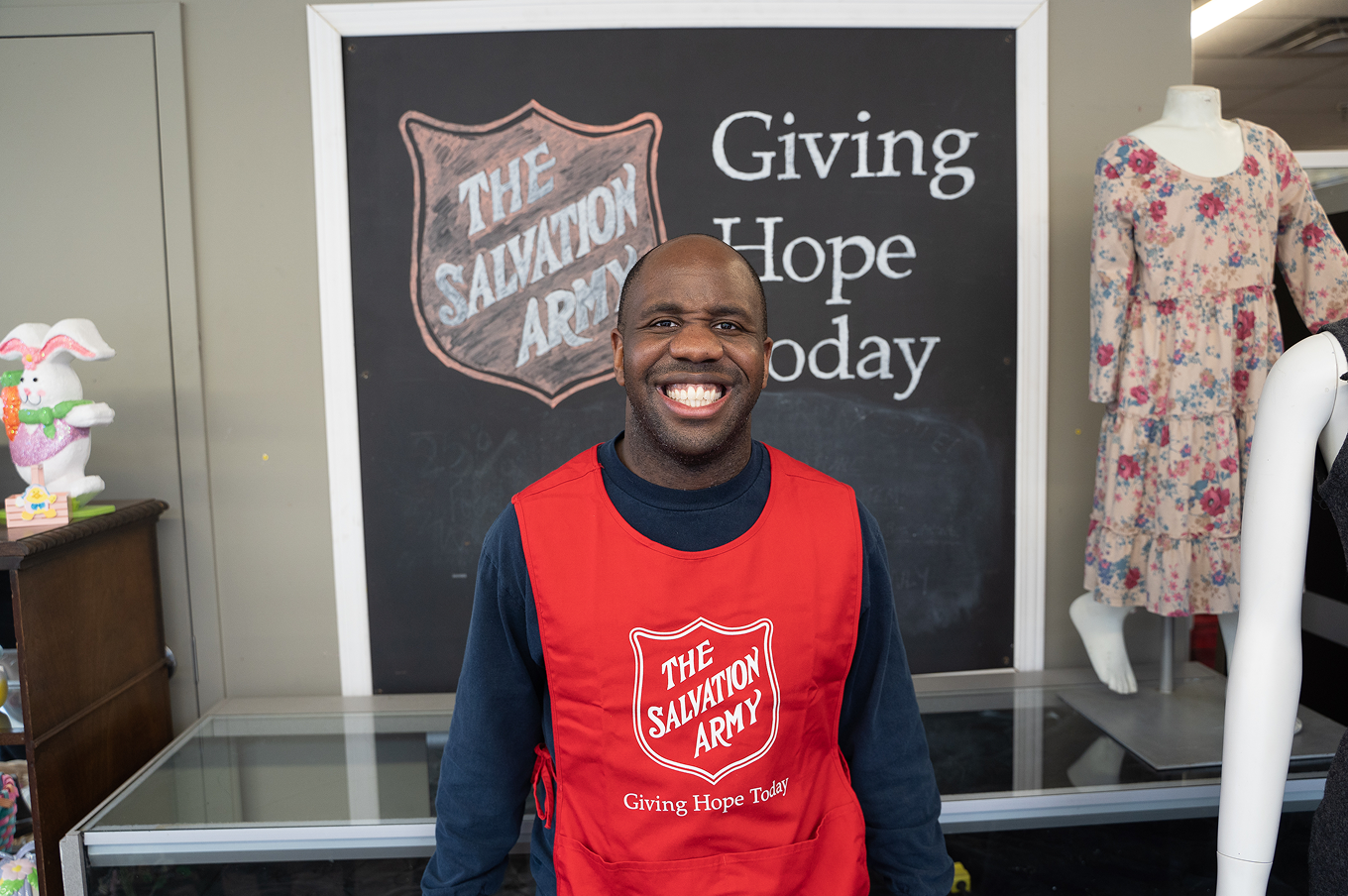 Smiling Salvation Army staff member wearing a red apron stands in front of a Giving Hope Today sign.